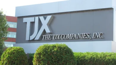 Outdoor sign for The TJX Companies, Inc. with large silver letters on a grey background, framed by green bushes.
